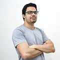 Man in a grey shirt standing, looking directly at the camera with a neutral expression.