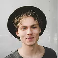A man wearing a stylish hat, standing outdoors with a neutral expression on his face.